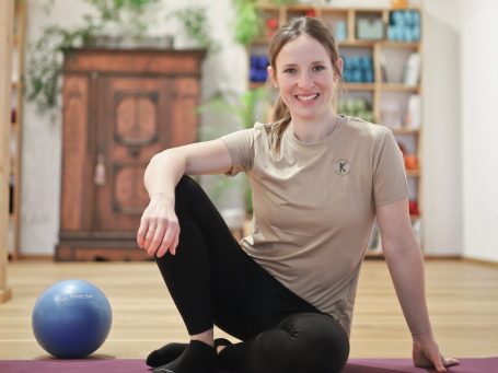 Pilatestrainerin lächelt in die Kamera, sitzt leicht seitlich und hat einen Ball vor sich liegen